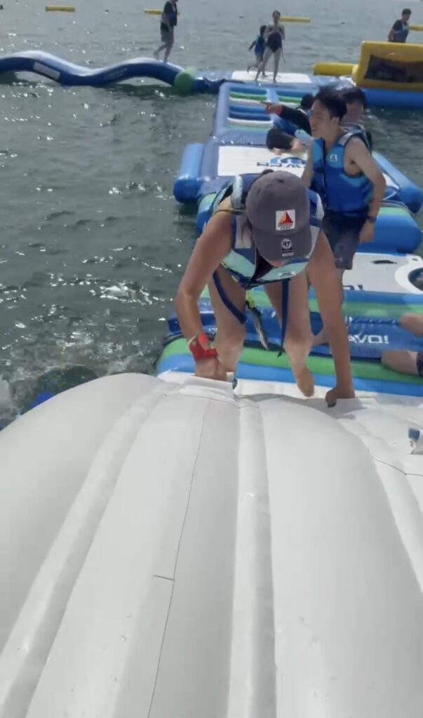 逗子海岸ウォーターパークの巨大な白い浮き遊具に、子どもが両手を使ってよじ登っている様子。後ろには海と他の参加者が見える。