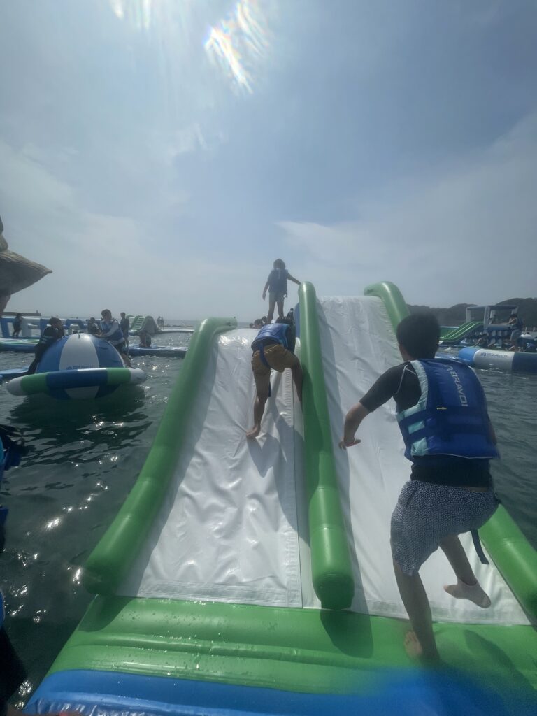 逗子海岸ウォーターパークの緑と白の大型スロープを、ライフジャケットを着た子どもと大人が登っている様子。