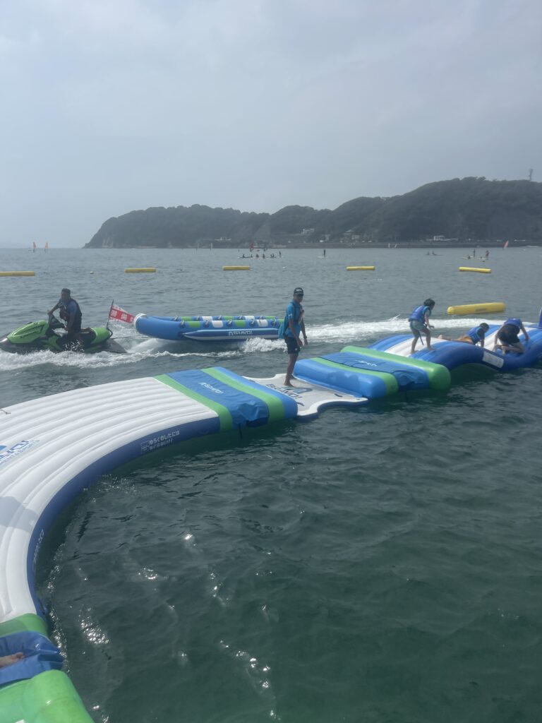 神奈川県逗子海岸のウォーターパークで、青と緑の大型フロート上を歩く人たち。周囲は海に囲まれており、夏のマリンアクティビティを楽しんでいる様子。