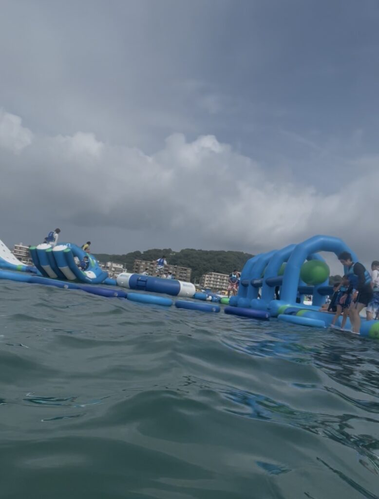 逗子海岸ウォーターパークを海上から見た様子。青と白の大型海上アスレチックが波間に浮かび、背景には曇り空と山並みが広がる。
