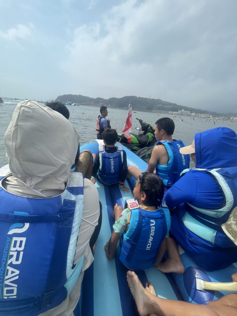 神奈川県逗子海岸のウォーターパークで、青いライフジャケットを着た子どもと大人が、バナナボートに乗って海上を進んでいる様子