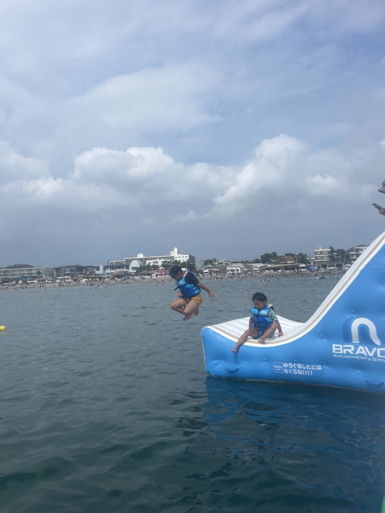 逗子海岸ウォーターパークの青いスライダーを滑る子どもと大人。海上アスレチックの一部で、夏の海遊びを楽しむ様子。