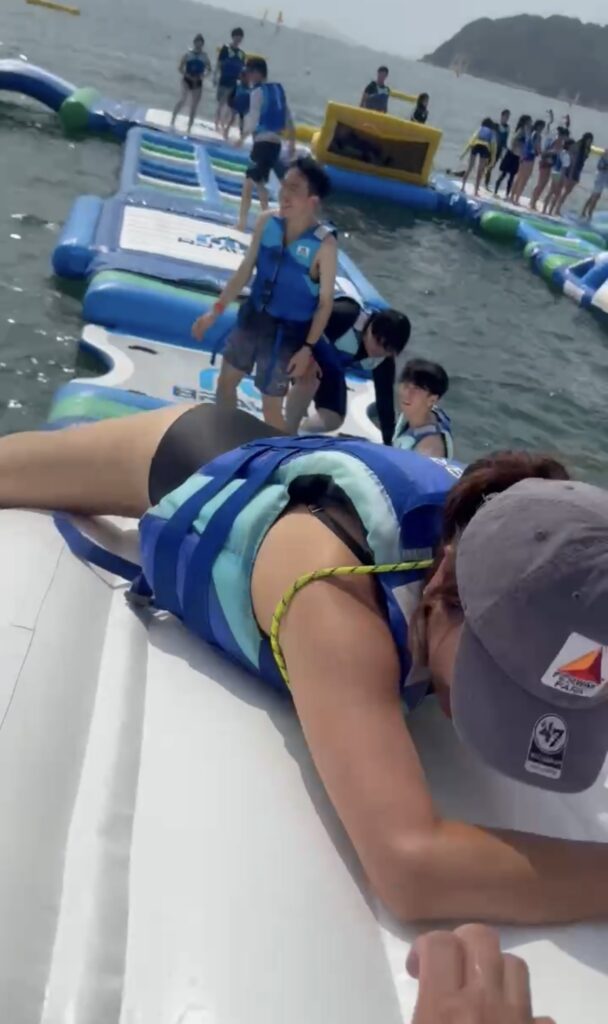 逗子海岸ウォーターパークの白い大型浮き具の上で、バランスを崩して落ちそうになっている様子。後方には海と他の利用者が見える。